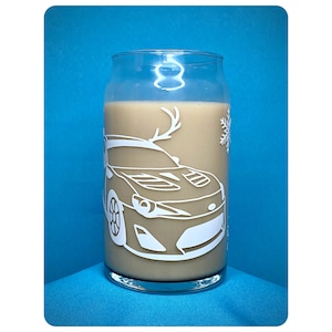 Puede incluir: Una taza de vidrio con un contorno blanco de un coche y un diseño de copo de nieve. La taza está llena de una bebida marrón claro.