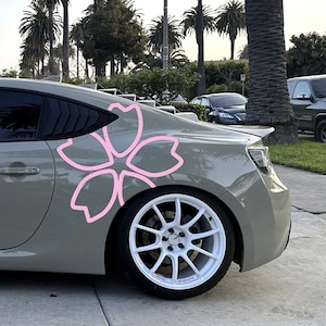 May include: A gray sports car with a large pink cherry blossom decal on the side. The car has white wheels and black tires. The background includes palm trees and other vehicles.