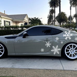 Può includere: Un'auto sportiva grigia con decalcomanie di fiocchi di neve bianchi sul lato. L'auto ha ruote bianche e vetri oscurati. Il veicolo è parcheggiato su una superficie pavimentata.