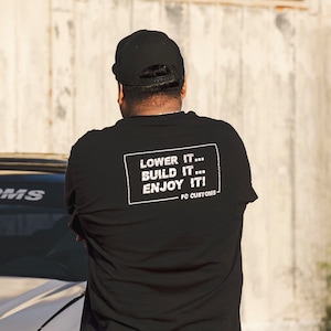 May include: Black long-sleeve shirt with the text "LOWER IT... BUILD IT... ENJOY IT!" in a white rectangular box. The shirt also has "FC CUSTOMS" printed below the text. The person is wearing a black cap.