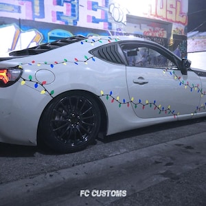 May include: A white car with a string of colorful Christmas lights on the side. The car is parked on a street with graffiti on the wall behind it.