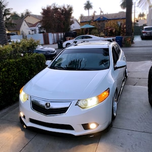 Puede incluir: Un Acura TL blanco con un parachoques delantero negro y un portaequipajes de techo negro. El coche está aparcado en una entrada de vehículos con una casa al fondo.