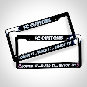 以下が含まれることがあります： 「FC CUSTOMS」と「LOWER IT...BUILD IT...ENJOY IT!」の銀色の文字が書かれた黒いナンバープレートフレーム2つ。下のフレームにはピンクの花のアクセントがある。