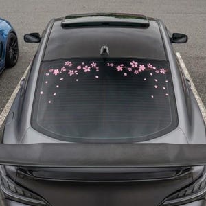 Puede incluir: Un coche gris oscuro con un adhesivo en la luneta trasera de flores de cerezo rosas y pétalos dispuestos en un patrón de rama horizontal en el borde superior, sobre un alerón trasero negro y un panel de techo negro.