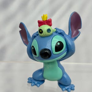 May include: A blue and green Stitch figurine with a small green alien wearing a red bow and yellow crown on its head.