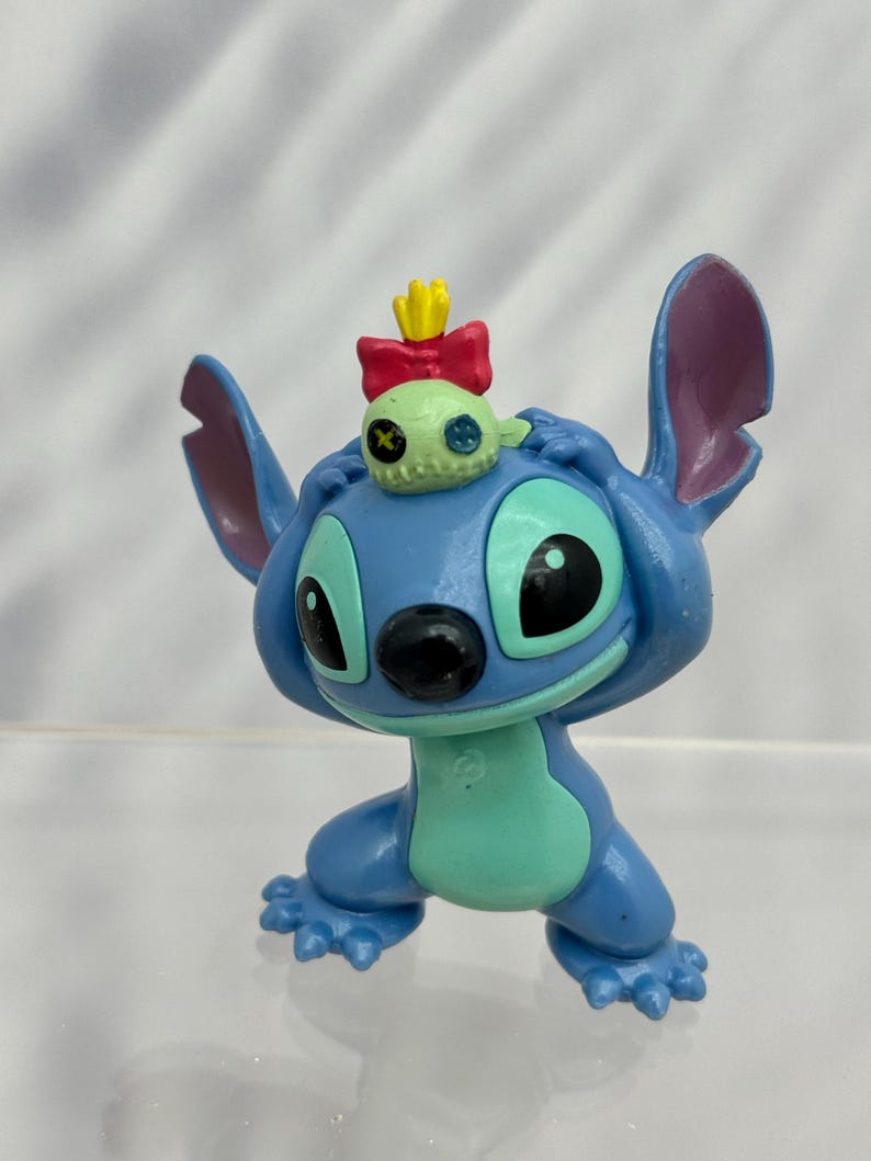 May include: A blue and green Stitch figurine with a small green alien wearing a red bow and yellow crown on its head.