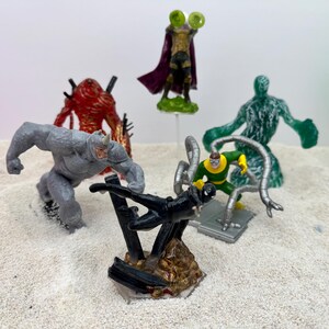 May include: A collection of superhero action figures, including Rhino, Mysterio, and Doctor Octopus. The figures are in various poses and colors, set against a sandy background. The figures are approximately 3-4 inches tall.