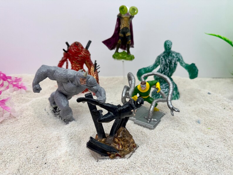 May include: A collection of superhero action figures, including Rhino, Mysterio, and Doctor Octopus. The figures are in various poses and colors, set against a sandy background. The figures are approximately 3-4 inches tall.