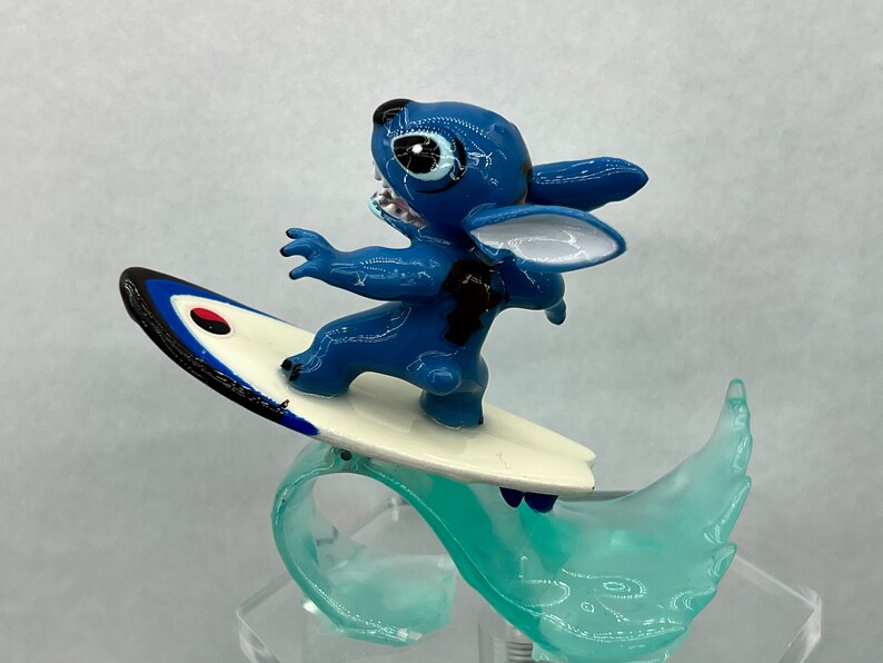Pu&ograve; includere: Una statuina blu di Stitch che cavalca un surf su un'onda turchese. La tavola da surf ha un design bianco e nero con un cerchio rosso.