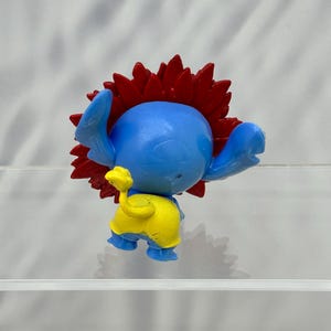 May include: Blue and yellow plastic toy figure with red spiky hair. The figure is standing on a clear surface.