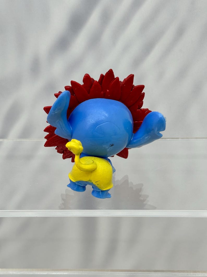 May include: Blue and yellow plastic toy figure with red spiky hair. The figure is standing on a clear surface.