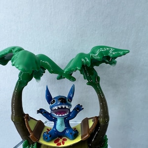 Pu&ograve; includere: Una statuina blu del personaggio Stitch seduto su un'amaca tra due palme verdi. L'amaca &egrave; marrone con una base gialla e un motivo a fiori rossi.