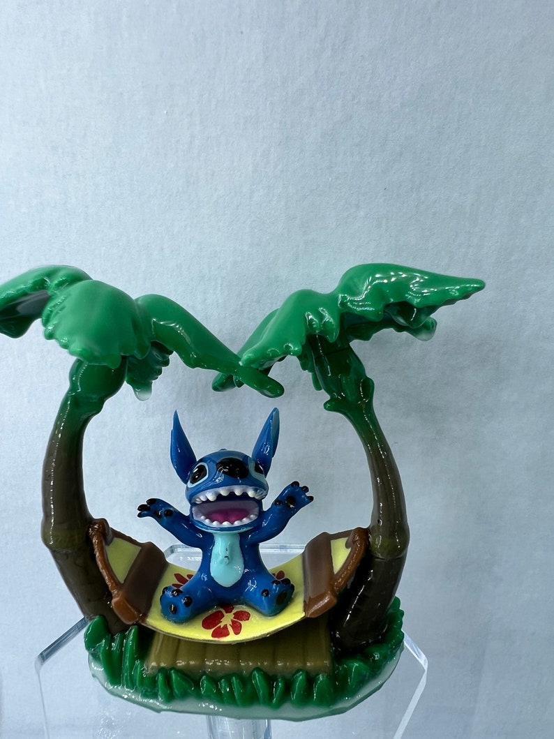 Pu&ograve; includere: Una statuina blu del personaggio Stitch seduto su un'amaca tra due palme verdi. L'amaca &egrave; marrone con una base gialla e un motivo a fiori rossi.