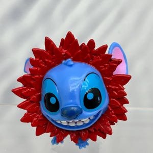 May include: Blue Stitch character with a red lion mane. The character has a big smile and large eyes.
