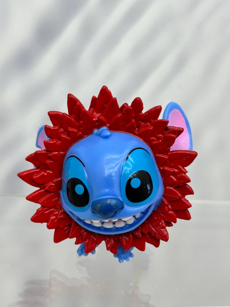 May include: Blue Stitch character with a red lion mane. The character has a big smile and large eyes.