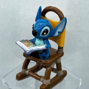 Pu&ograve; includere: Una statuina blu di Stitch seduta su una sedia a dondolo, che legge un libro. La sedia &egrave; marrone con accenti gialli.
