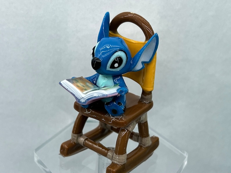 Pu&ograve; includere: Una statuina blu di Stitch seduta su una sedia a dondolo, che legge un libro. La sedia &egrave; marrone con accenti gialli.