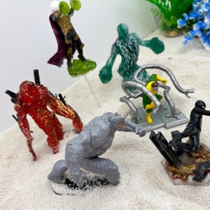 May include: A collection of colorful action figures featuring various characters. The figures include a red monster, a gray rhino-like figure, a green figure, a figure with metal arms, a black figure, and a figure with a purple cape.
