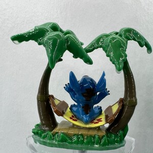Pu&ograve; includere: Una statuina blu del personaggio Stitch che si rilassa in un'amaca tra due palme verdi. La statuina &egrave; seduta su un'amaca gialla e marrone con fiori rossi. La statuina &egrave; su una base verde con un supporto in acrilico trasparente.