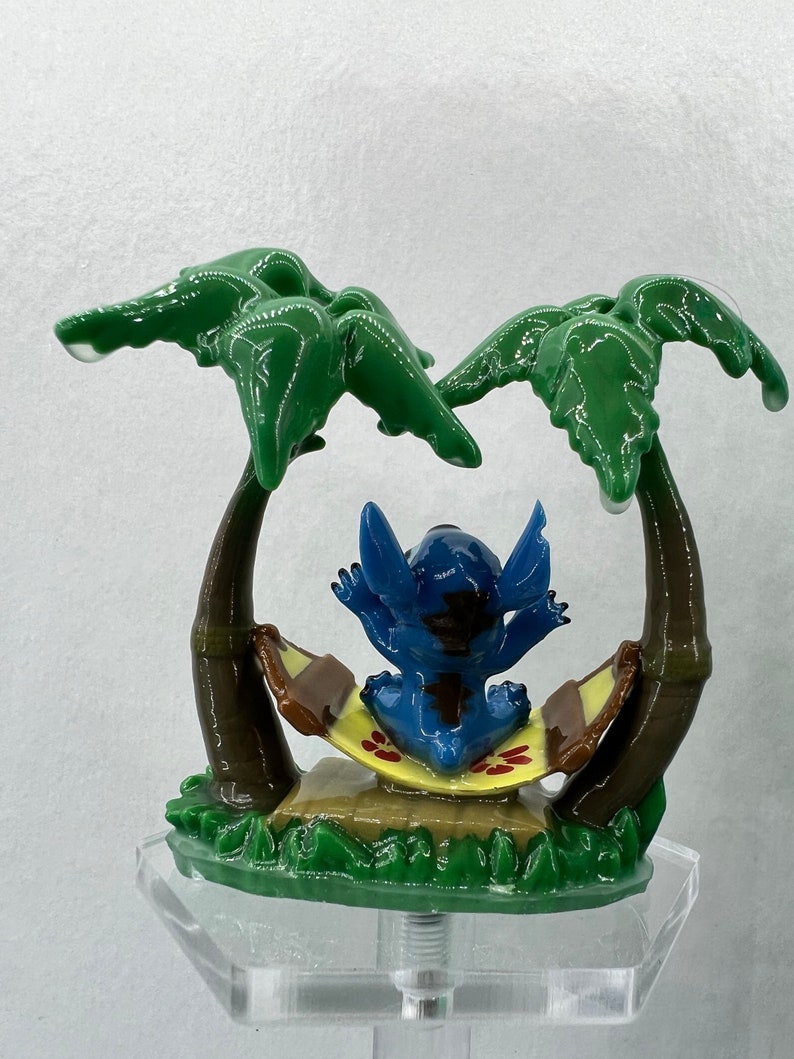 Pu&ograve; includere: Una statuina blu del personaggio Stitch che si rilassa in un'amaca tra due palme verdi. La statuina &egrave; seduta su un'amaca gialla e marrone con fiori rossi. La statuina &egrave; su una base verde con un supporto in acrilico trasparente.