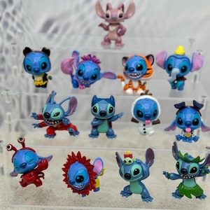 May include: A collection of 14 blue Stitch figurines in various poses and costumes. The figurines are displayed on a clear acrylic shelf. Some of the costumes include a tiger, a snowman, a crab, and a hula dancer.
