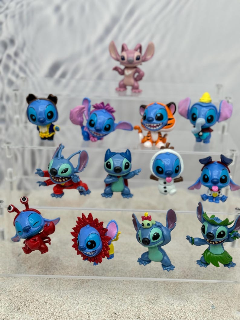 May include: A collection of 14 blue Stitch figurines in various poses and costumes. The figurines are displayed on a clear acrylic shelf. Some of the costumes include a tiger, a snowman, a crab, and a hula dancer.