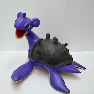 May include: A purple Lapras figure with a black shell. The toy has a long neck, flippers, and a smiling face with a red mouth. The figure is a collectible item.