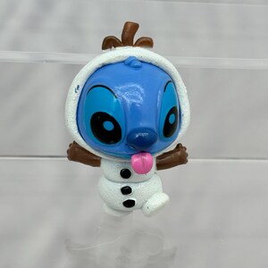 May include: A blue Stitch character figurine dressed as a snowman. The figurine has a white snowman costume with black buttons and a pink tongue sticking out. The figurine has brown arms and a brown hat.