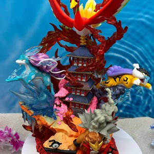 Puede incluir: Figura detallada que presenta una criatura con forma de ave roja, amarilla y blanca con las alas extendidas sobre una pagoda. También se representan otras criaturas en tonos azules, morados y amarillos, con elementos de fuego y hielo. La base es de color naranja fuego.