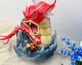 Può includere: Una figura di Gyarados rossa e crema con la bocca aperta, ambientata in una base a tema marino con una figura di Magikarp più piccola. La figura è dettagliata con una posa dinamica ed è circondata da effetti d'acqua blu e fiori artificiali.