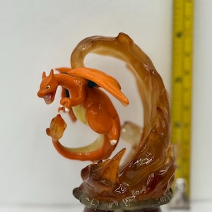 May include: A detailed figurine of Charizard, a Pokémon character, in a dynamic pose. The figure is primarily orange with blue accents on its wings. It is surrounded by a stylized flame effect, with a brown and amber color scheme. The base is a dark brown.