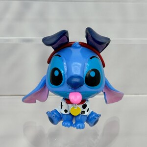 May include: A blue Stitch figurine wearing a red bandana and a white and black spotted shirt. Stitch is sticking out its pink tongue and has a yellow collar with a tag.
