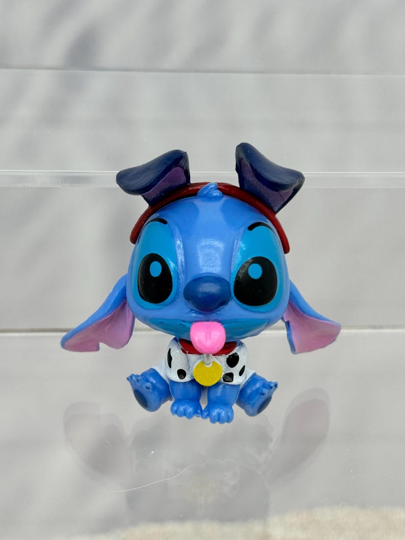 May include: A blue Stitch figurine wearing a red bandana and a white and black spotted shirt. Stitch is sticking out its pink tongue and has a yellow collar with a tag.