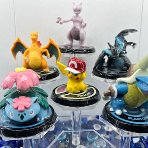 May include: A set of six Pokemon figures on black bases with the names "Blastoise", "Pikachu", and "Charizard" printed on them. The figures are arranged in a circle on a clear acrylic stand.