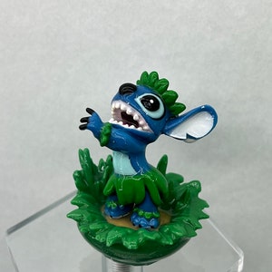 Pu&ograve; includere: Una statuina blu di Stitch con un lei verde e foglie verdi intorno alla base. La statuina sorride a bocca aperta e con gli occhi spalancati.