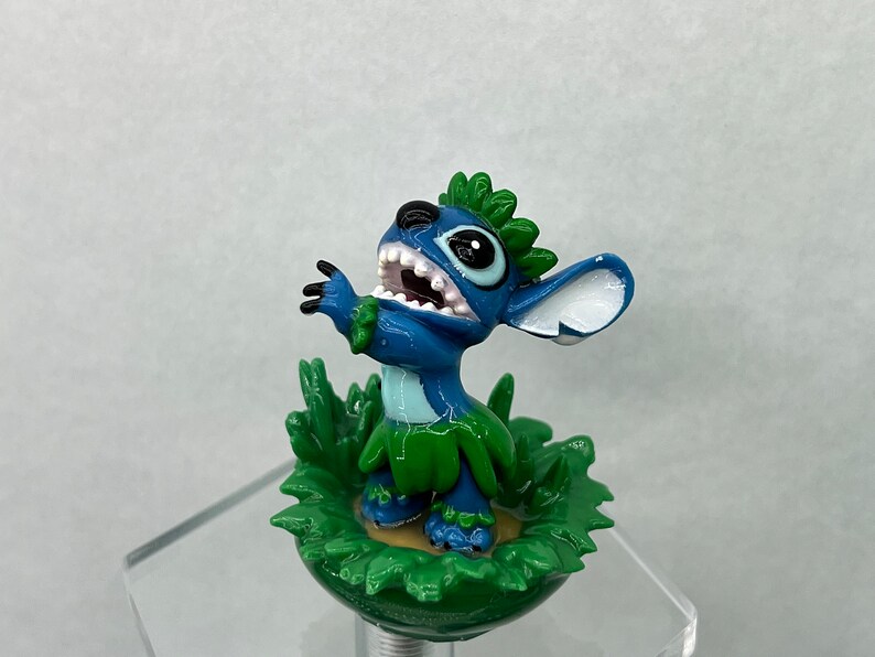 Pu&ograve; includere: Una statuina blu di Stitch con un lei verde e foglie verdi intorno alla base. La statuina sorride a bocca aperta e con gli occhi spalancati.