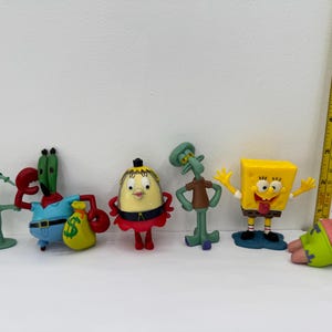 Könnte beinhalten: Eine Sammlung von sechs farbenfrohen Figuren der SpongeBob Schwammkopf-Charaktere. Die Figuren umfassen Plankton, Mr. Krabs, Pearl, Thaddäus, SpongeBob und Patrick. Ein gelbes Lineal befindet sich vertikal im Hintergrund.
