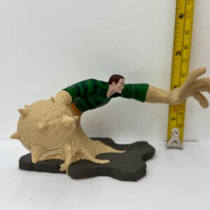 May include: A Sandman action figure, featuring a green and tan striped shirt, tan arms, and a sand-like base. The figure is posed with one arm extended, and a spiked ball-shaped structure. A yellow ruler is next to the figure.