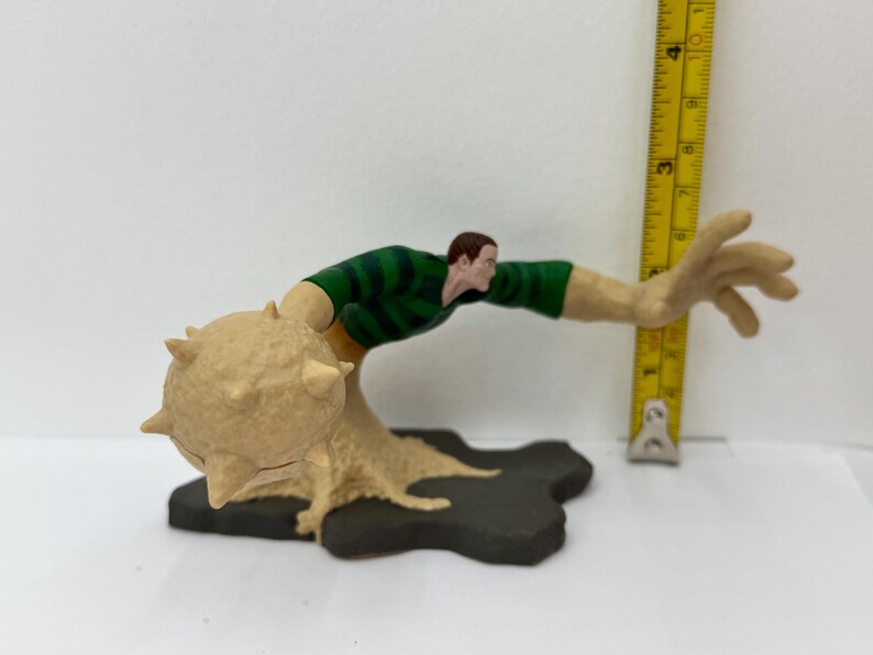 May include: A Sandman action figure, featuring a green and tan striped shirt, tan arms, and a sand-like base. The figure is posed with one arm extended, and a spiked ball-shaped structure. A yellow ruler is next to the figure.
