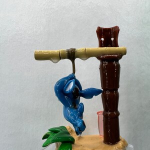 Pu&ograve; includere: Una statuina blu di Stitch appesa a un palo di bamb&ugrave; marrone con foglie verdi alla base. La statuina &egrave; sospesa da un palo di legno marrone con una tazza rosa.
