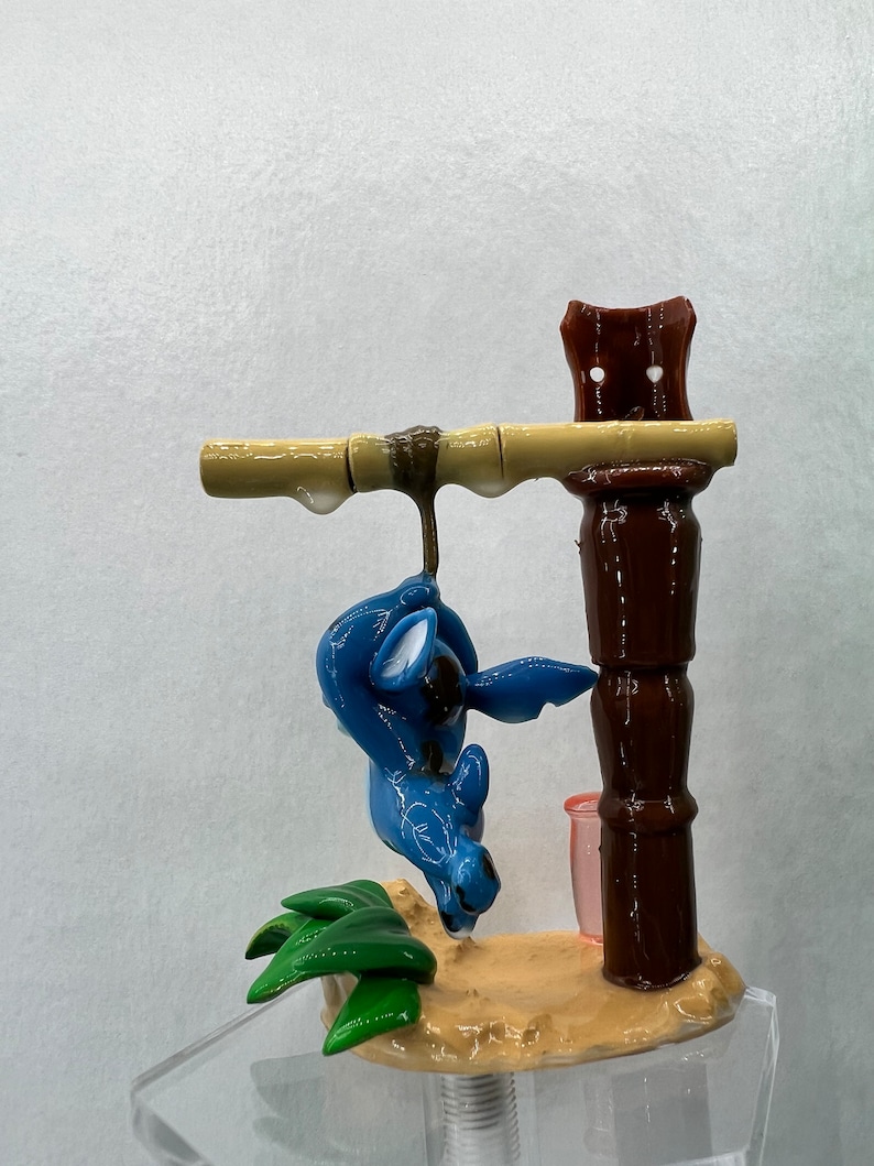Pu&ograve; includere: Una statuina blu di Stitch appesa a un palo di bamb&ugrave; marrone con foglie verdi alla base. La statuina &egrave; sospesa da un palo di legno marrone con una tazza rosa.