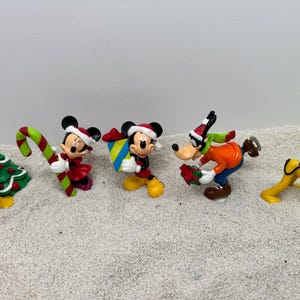 May include: A set of six Christmas-themed figurines featuring Mickey Mouse, Minnie Mouse, Pluto, and Goofy. The figurines are made of plastic and are painted in bright colors. Mickey and Minnie are wearing Santa hats, Pluto is holding a candy cane, and Goofy is wearing a scarf and carrying a gift.