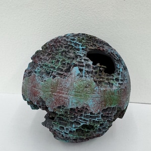 May include: A textured, spherical decorative object with a dark interior. The exterior features a rough, porous surface with a blend of colors, including teal, burgundy, and green. The object has a unique, weathered appearance.