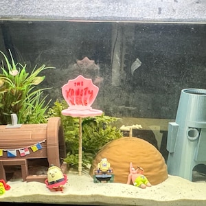 May include: A decorative aquarium scene featuring various SpongeBob SquarePants themed ornaments. The scene includes a bucket with "Krusty Krab" text, a treasure chest, a pineapple house, and other character figurines, all set against a backdrop of artificial plants.