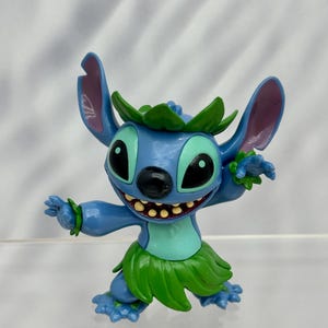 May include: A blue and green Stitch figurine wearing a lei and a grass skirt. The figurine is in a dancing pose with one arm raised and the other arm bent at the elbow.