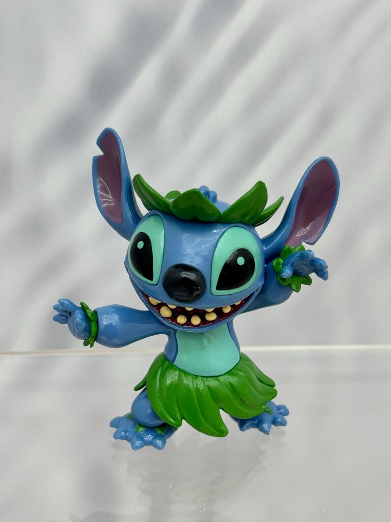 May include: A blue and green Stitch figurine wearing a lei and a grass skirt. The figurine is in a dancing pose with one arm raised and the other arm bent at the elbow.
