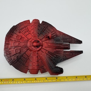 May include: A red and black spaceship model with a circular main body and a forward-facing cockpit section. A yellow measuring tape is positioned along the bottom edge, showing the object spans approximately 8.5 inches in total width.