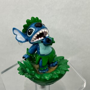 Pu&ograve; includere: Figurina di Stitch blu con capelli verdi e gonna d'erba, in piedi su una base di erba verde. Il personaggio ha un'espressione sorpresa con la bocca aperta e una singola lacrima sulla guancia.