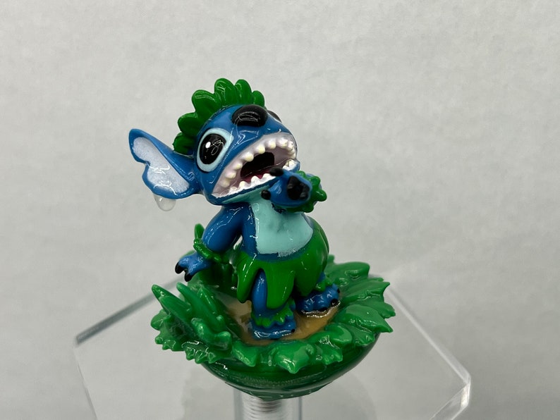 Pu&ograve; includere: Figurina di Stitch blu con capelli verdi e gonna d'erba, in piedi su una base di erba verde. Il personaggio ha un'espressione sorpresa con la bocca aperta e una singola lacrima sulla guancia.