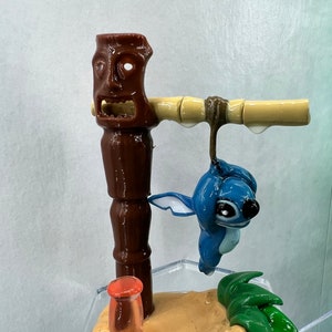 Pu&ograve; includere: Una statuina blu del personaggio Stitch appesa a un totem tiki marrone con un palo di bamb&ugrave;. La statuina &egrave; sospesa sopra una base sabbiosa con foglie verdi e un tubo di vetro rosso trasparente.